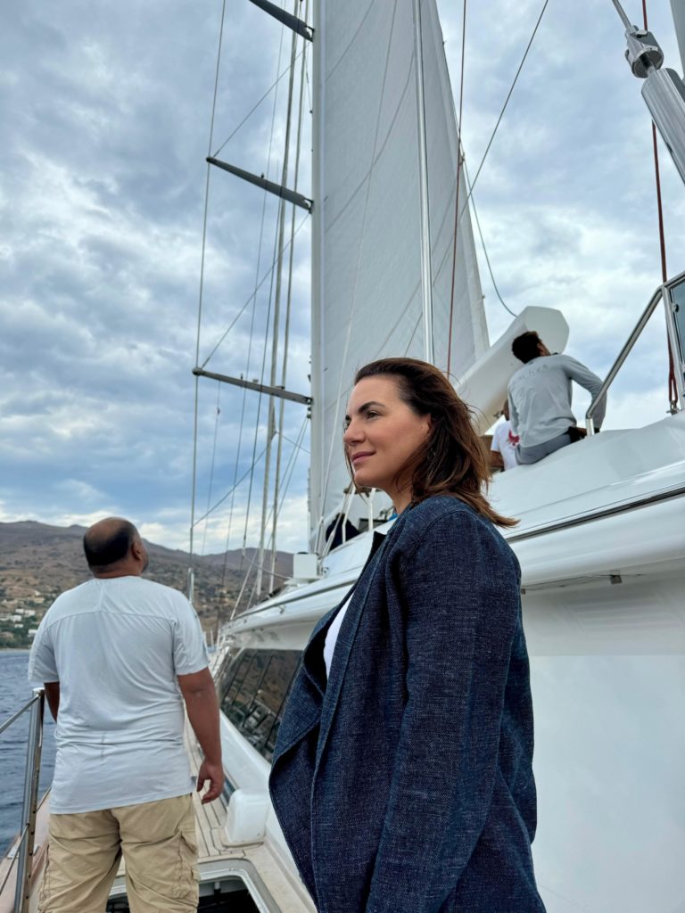 A woman stands on a sailboat, looking out at the water, while a man adjusts the sails in the background under a cloudy sky.