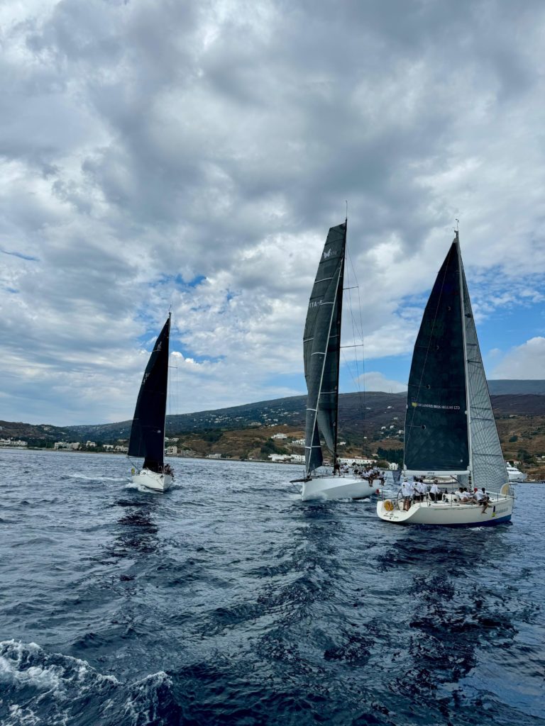 Three sailboats with black sails navigate through calm waters under a cloudy sky, with a hilly coastline in the background.