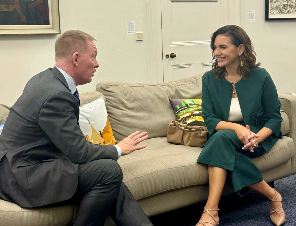 A man in a suit and a woman in a green outfit are engaged in conversation on a couch, with decorative pillows in the background.